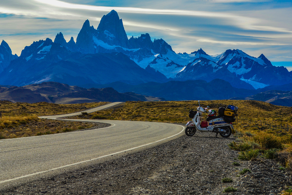 Vespa loaded with luggage on the side of a lonely winding road. Snow-covered mountains in the background.