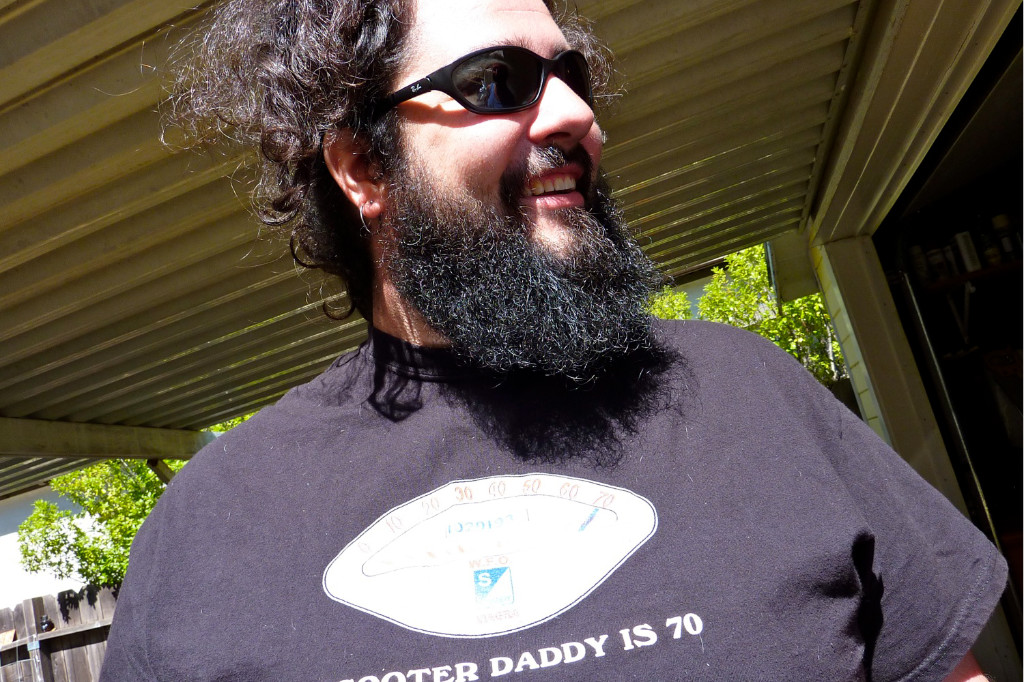 Ein lächelnder Mann mit dunklen Haaren und langem dunklen Bart, schwarzem T-Shirt und Sonnenbrille.