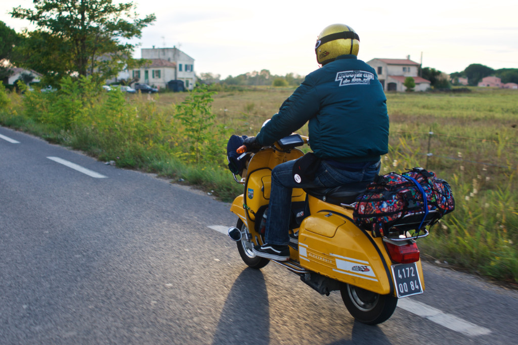 Eine Person fährt mit einer gelben Vespa die Landstraße entlang. Auf dem Gepäckträger befindet sich eine Tasche.