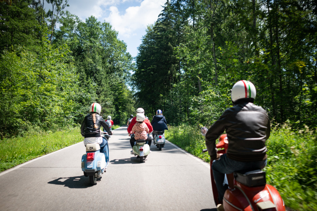 Several Vespa riders on a road. Forest to the left and right.