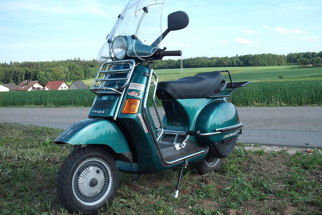 Vespa Cosa in dunkelgrün-metallic steht neben einer ländlichen Straße auf dem Rasen. Im HIntergrund sind einzelne Häuser zu erkennen.