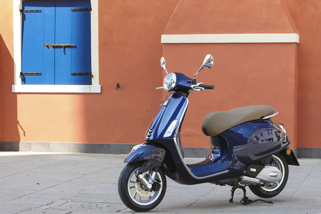 Blaue Vespa Primavera vor einer rotbraunen Hauswand und einem Fenster mit blauen Fensterläden