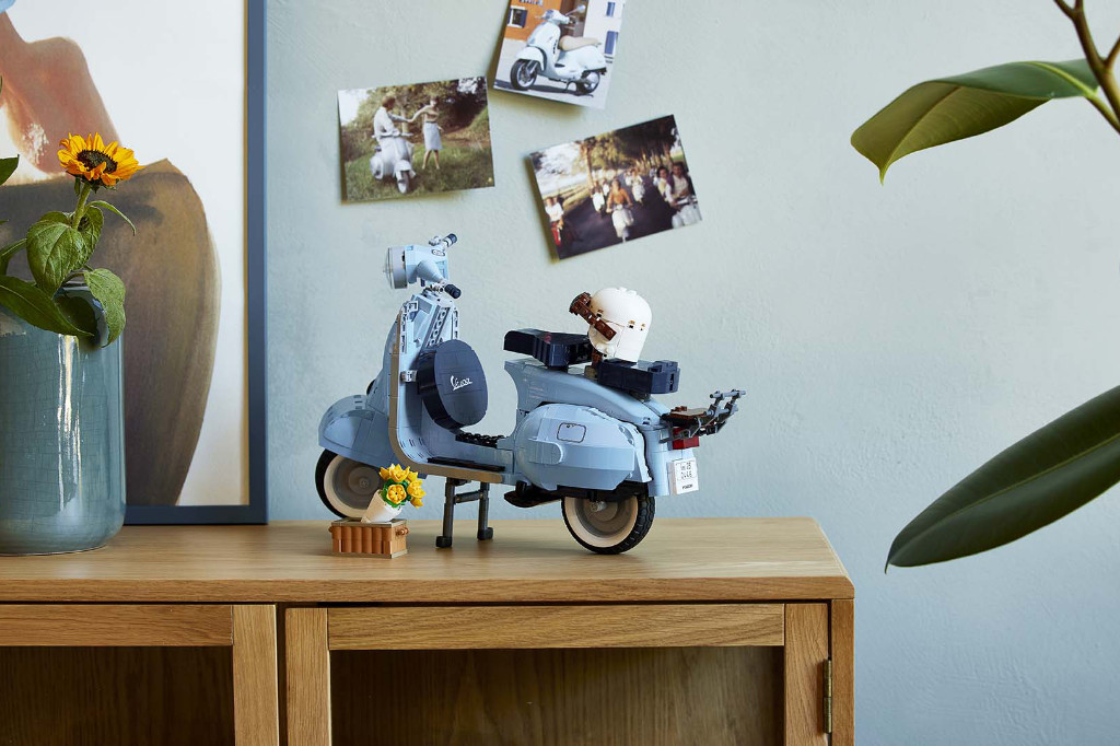 Die pastellblaue Lego Vespa steht vor einer hellen Wand auf einer Kommode. Darüber sind Fotos an der Wand angebracht, die Vespas und VespafahrerInnen zeigen. Links und rechts im Bild sind Pflanzen und ein Bilderrahmen zu erkennen.