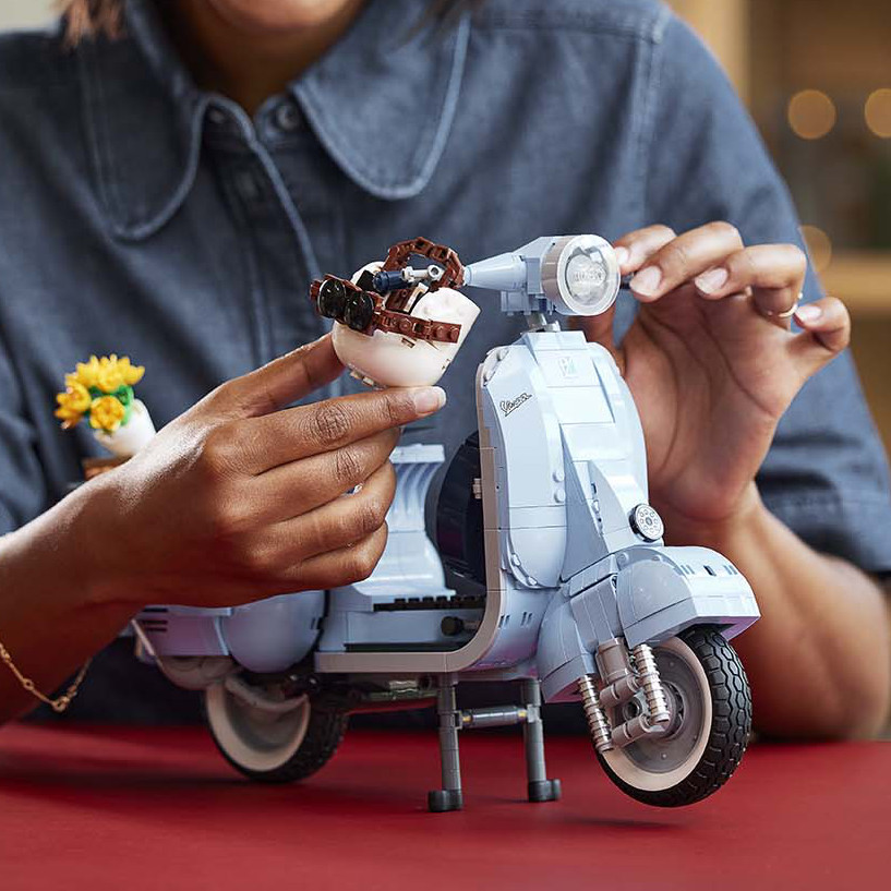 Die blaue Lego Vespa steht auf rotem Untergrund. Eine Person mit blauem Jeanshemd und Schmuck an den Händen sitzt dahinter und hängt den Helm an den Lenker der Vespa.