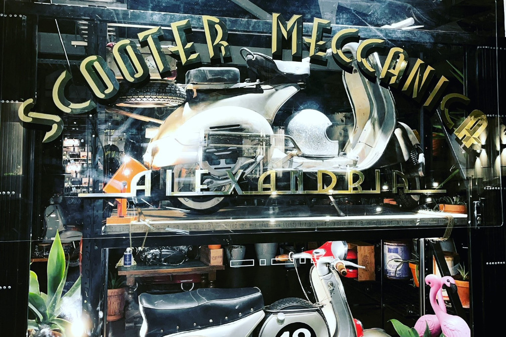 Scooter Meccanica Schriftzug auf einer Scheibe, hinter der ausgestellte Vespas zu sehen sind