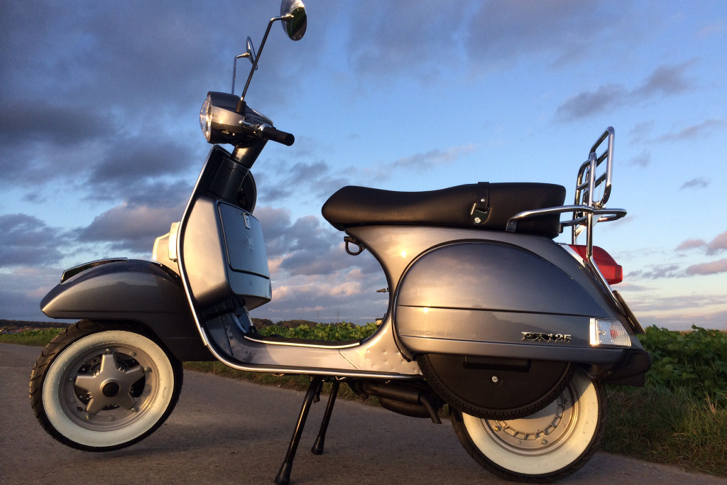 Metallic graue Vespa auf einer Straße und im Hintergrund bllauer Himmel mit Wolken.