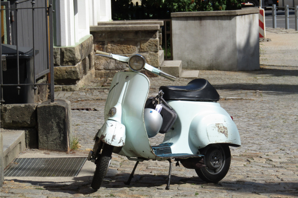 Helle blau-grüne Vespa 50 steht in einem Hof. Der Helm und eine Tasche hängen vorne an der Sitzbank.