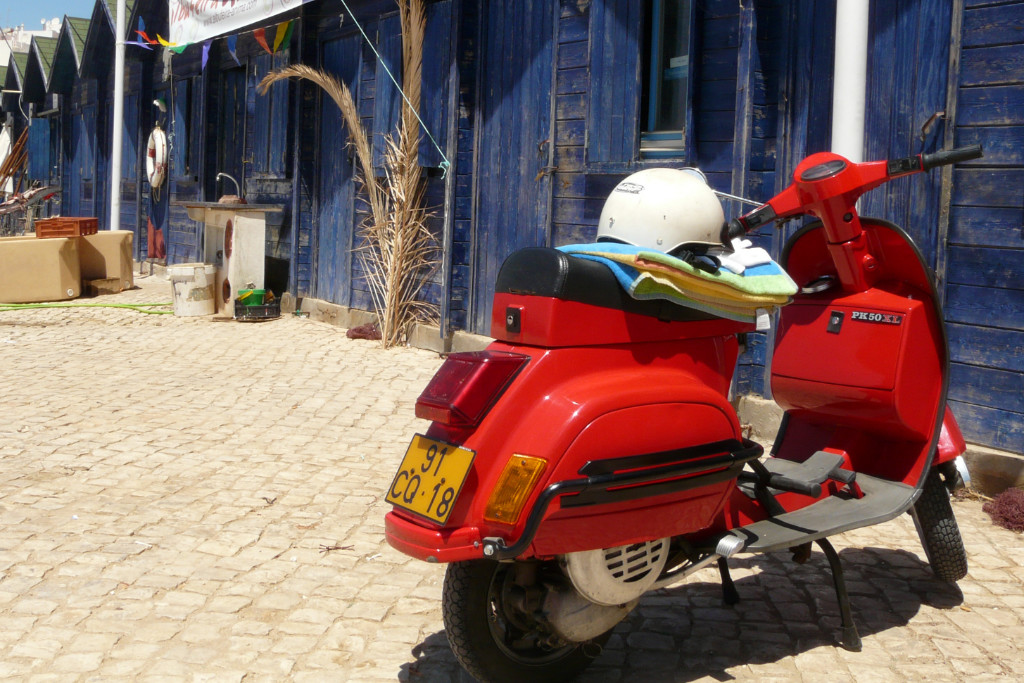 Rote Vespa mit einem zusammengelegten Handtuch und Helm auf der Sitzbank steht vor einem blauen Strandhaus aus Holz, an dem die FArbe schon etwas abblättert.