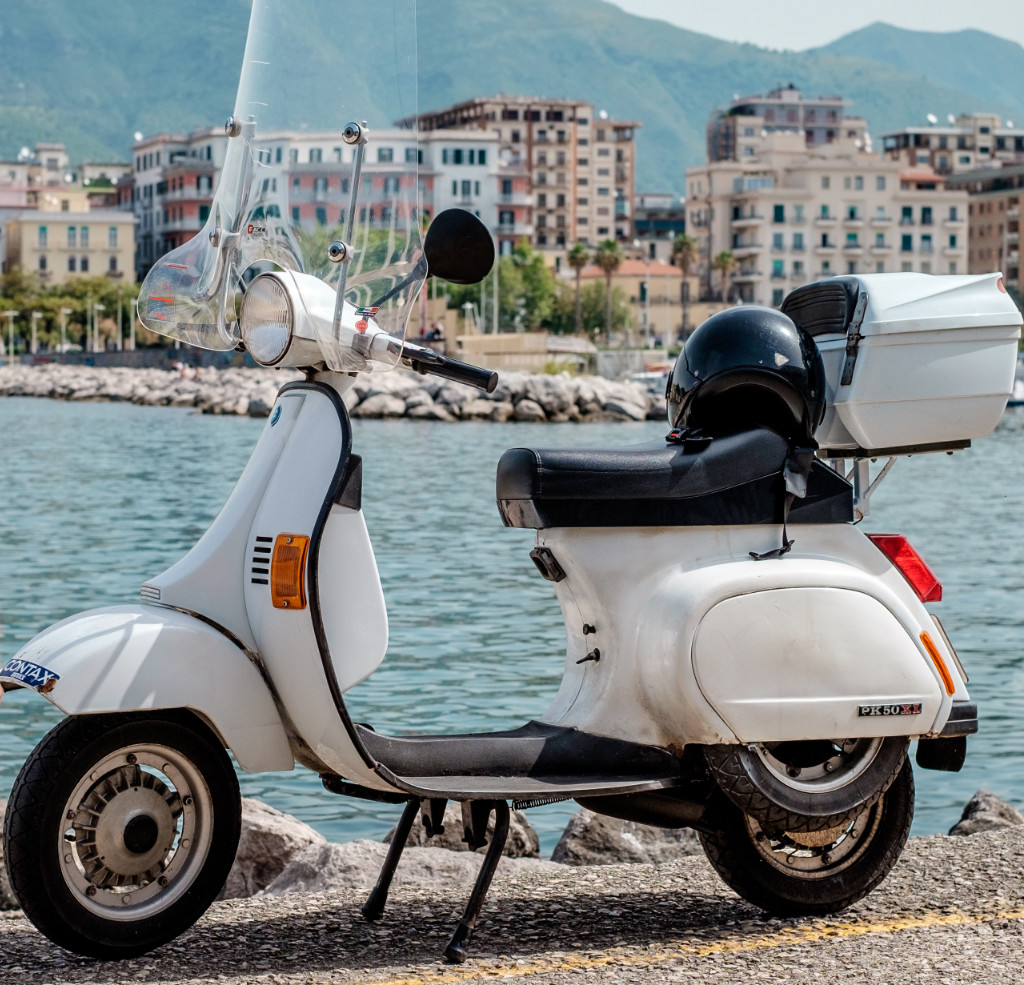 Weiße Vespa steht am Ufer eines Gewässers. Am anderen Ufer sind Häuser und Palmen zu erkennen.