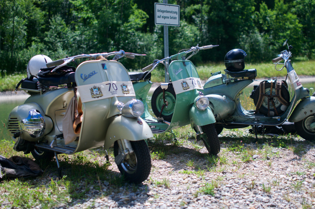 3 Wideframe Vespas stehen auf einer Wiese. Helme und anderes Zubehör liegen auf den Sitzbänken. Im Hintergrund sind Bäume zu sehen.