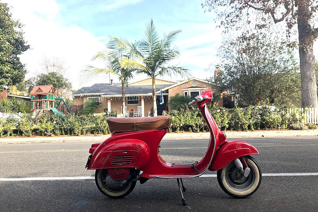 Rote Vespa 90 (restauriert) auf einer Straße. Im Hintergrund ein Haus und Palmen.