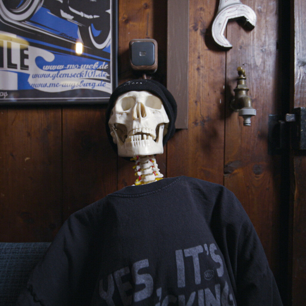 Skeleton with black jumper and black cap in front of a wooden wall with further decorative items.