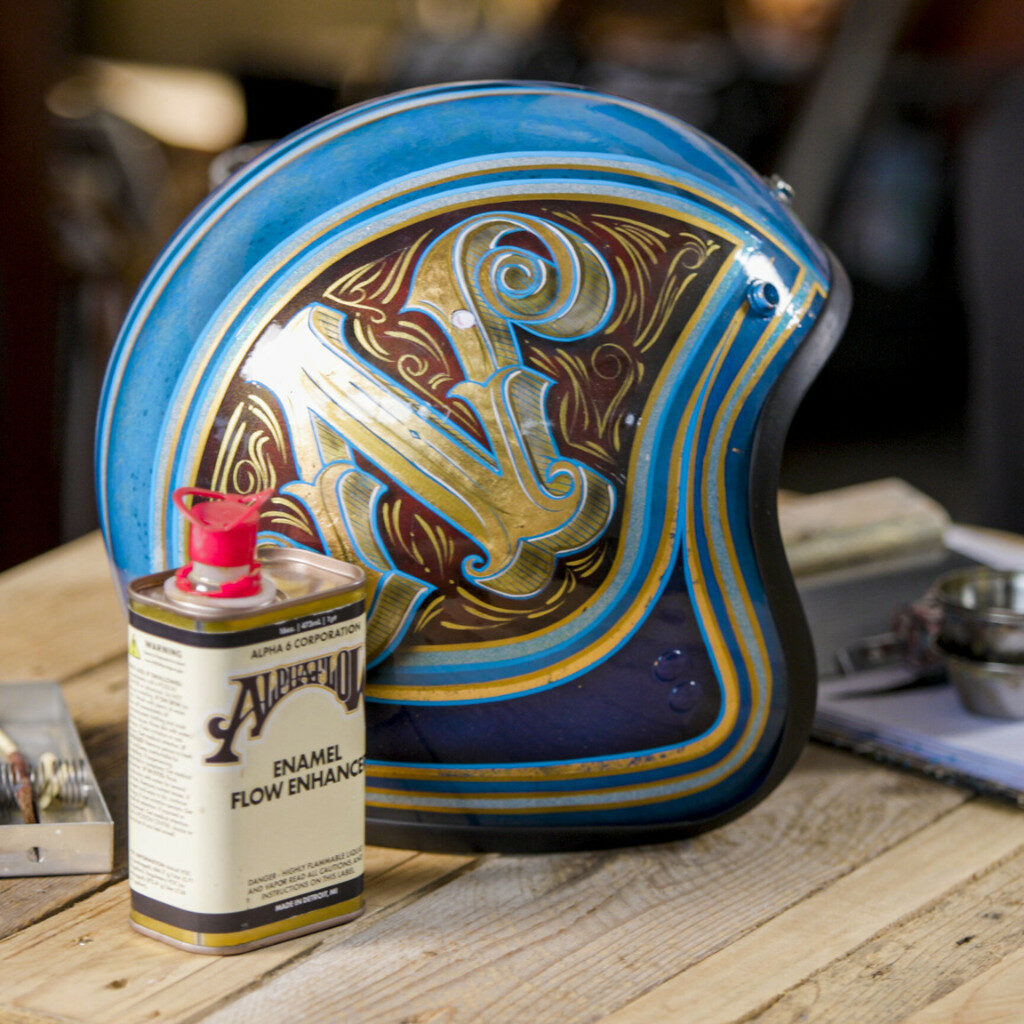 Blue and gold artfully decorated Vespa helmet on a table next to a bottle of "enamel flow enhancer