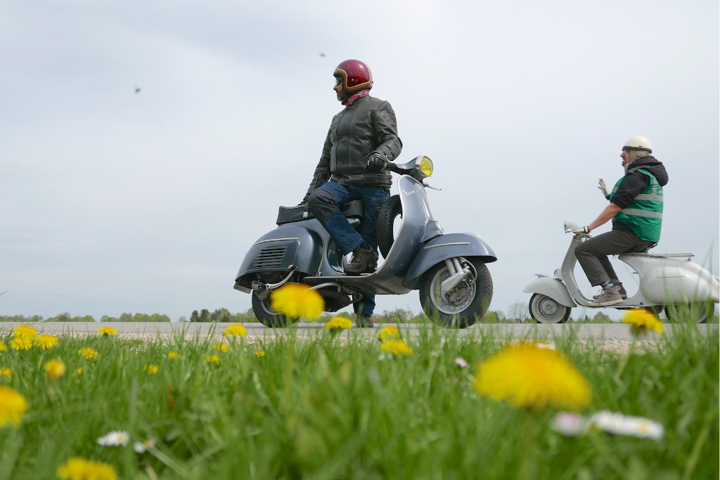 Two Vespa riders next to a meadow with flowering dandelions