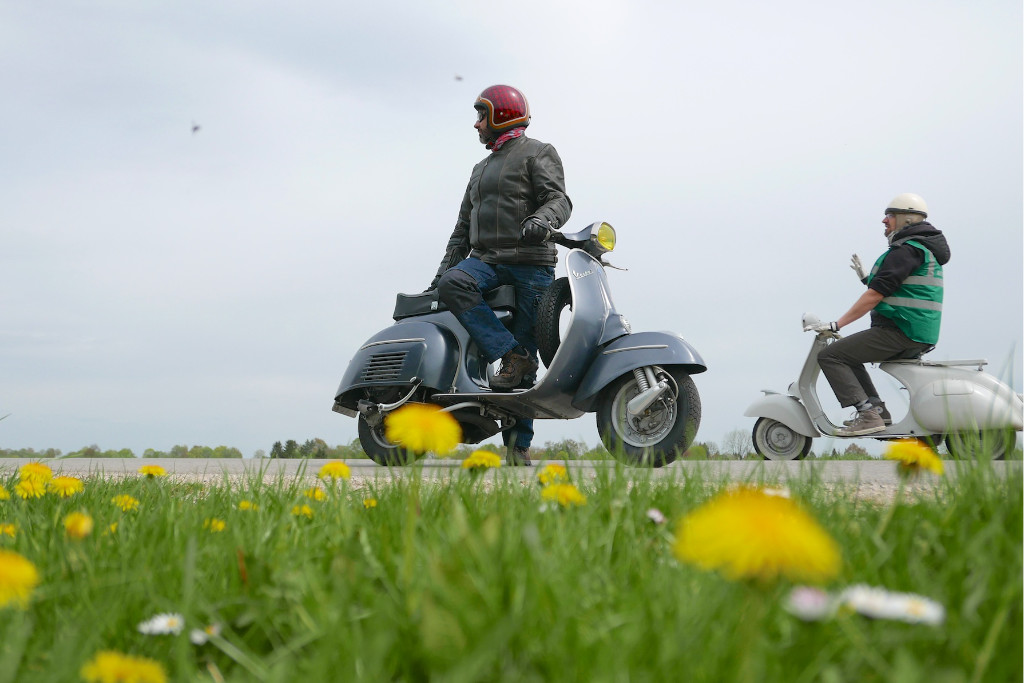 Zwei Vespa Fahrer stehen am Rand einer Wiese mit blühendem Löwenzahn