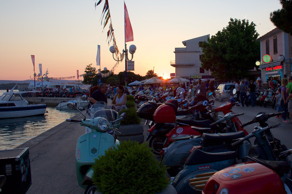 Viele Vespas stehen aufgereiht an einem belebten Hafen. Im Hintergrund geht die Sonne unter.