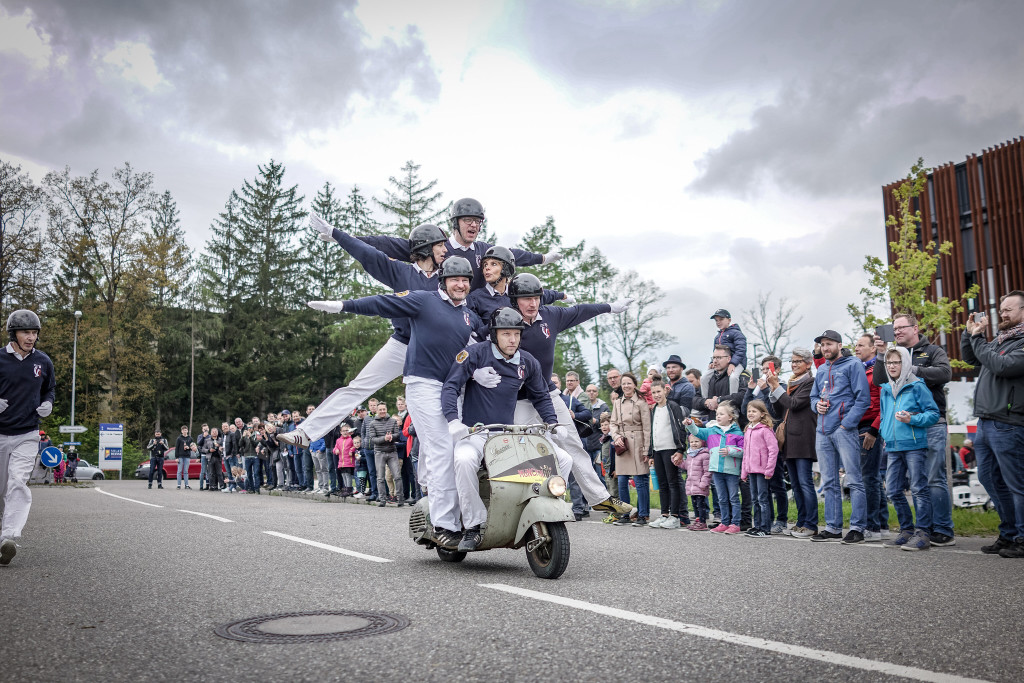 6 people perform an acrobatic show on an old Vespa. An enthusiastic audience stands on the sidelines.