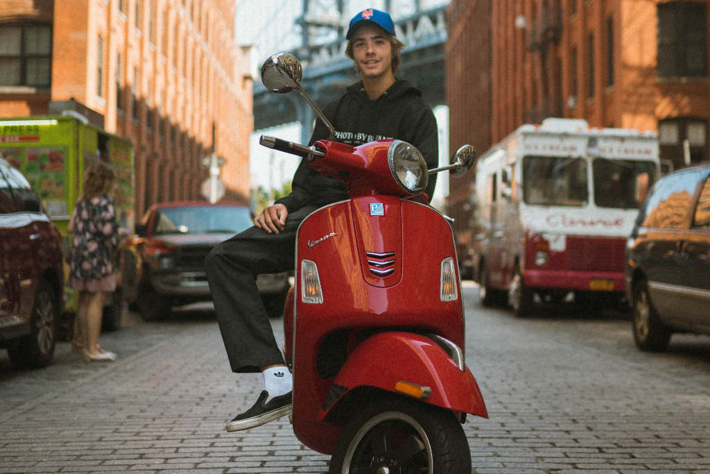 A teenager is sitting casually on a red Vespa in the middle of a small street in a city. Parked cars and brick buildings can be seen at the edge.