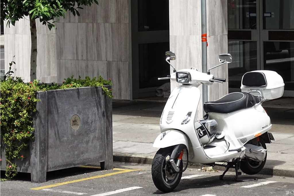 Eine weiße Vespa ist vor einem Gebäude auf einem Parkplatz für Krafträder geparkt. Daneben steht ein Baum in einem Container.
