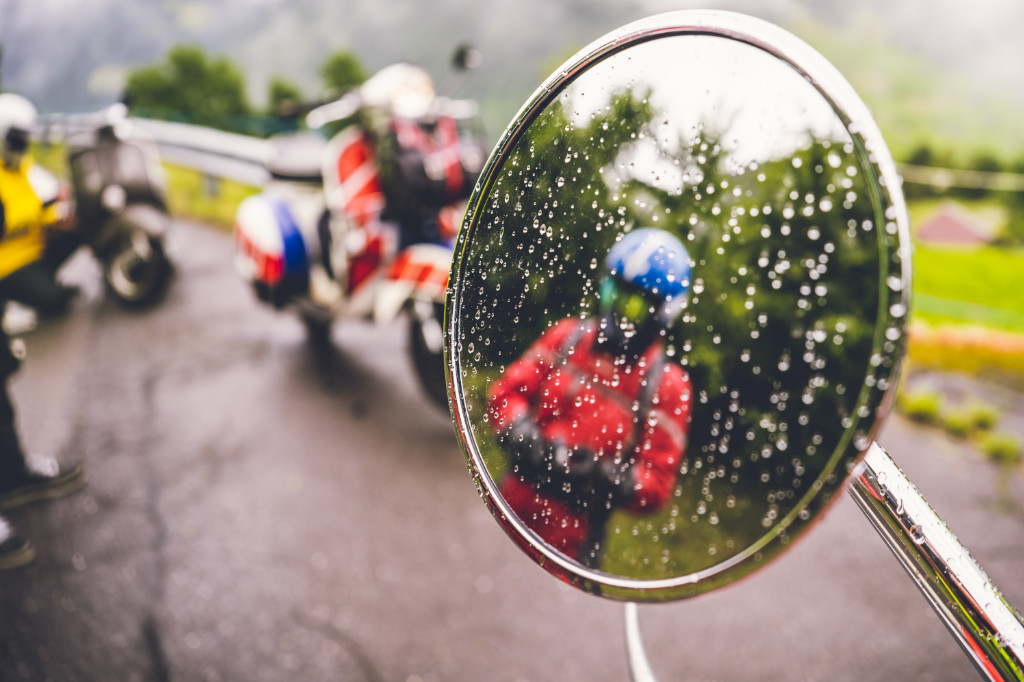 Vespa Spiegel mit Regentropfen, in dem sich andere Vespas und FahrerInnen auf einer Straße spiegeln