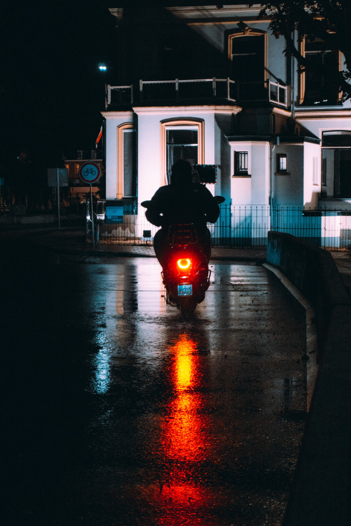 Eine Vespa bei Regen und Dunkelheit auf einer Straße vor einem beleuchteten weißen Haus