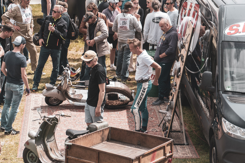 Several people standing around 2 exhibited old Vespas and other products on display in front of a transporter from SIP Scootershop