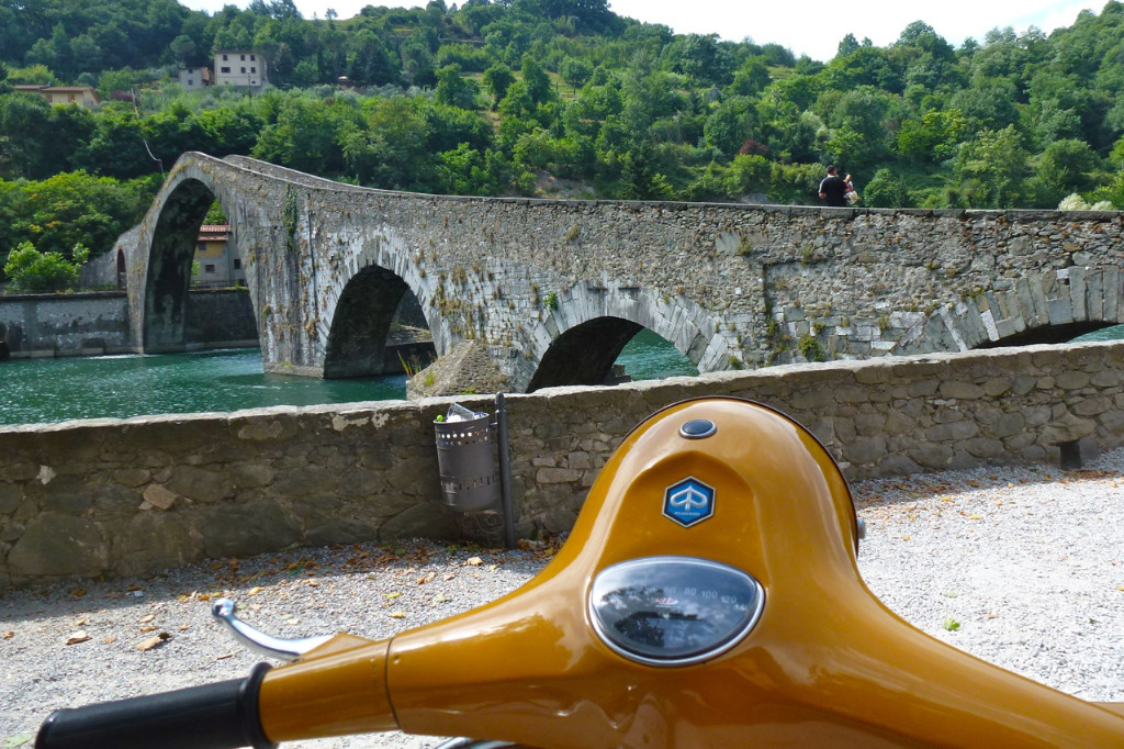 Im vordergrund ist der Ausschnitt eines gelben Vespa Lenkers zu sehen und im HIntergrund eine alte Steinbrücke, die über einen Fluss umgeben von Bäumen führt