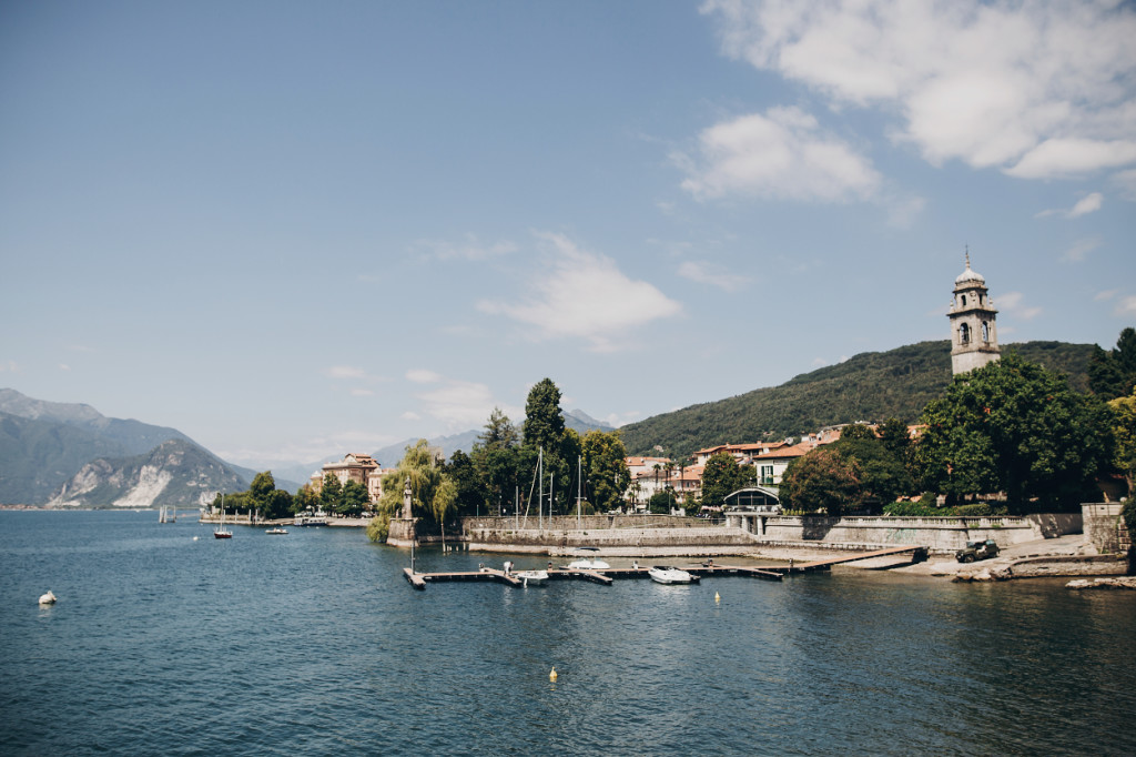 View of old buildings and boats on the lake in Stresa town, Italy. Architecture and shore on Lake Maggiore in sunny day on background of palm trees and mountains
