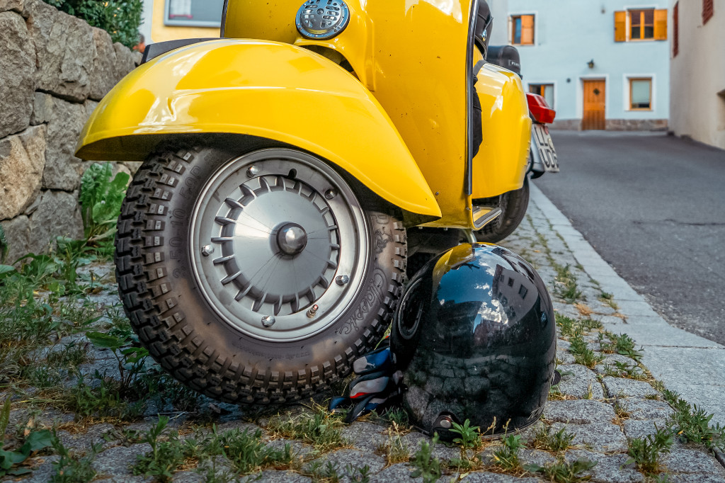 Vorderrad einer gelben Vespa, daneben ein Motorradhelm auf einer Straße