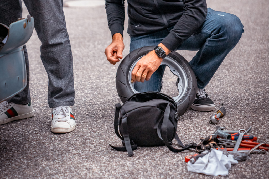 Eine Person kniet auf der Straße und hantiert an einem Reifen herum. daneben liegen eine Tasche und Werkzeug.